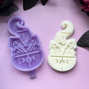 May include: Two silicone molds for resin casting, one purple and one white, shaped like a cauldron with a smoke plume and crystals. The cauldron has a moon and stars design.