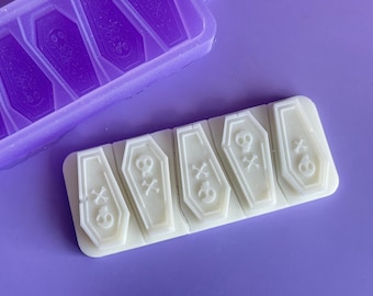 May include: A white silicone mold with six coffin-shaped cavities. Each cavity features a skull and crossbones design. The mold is on a purple surface.