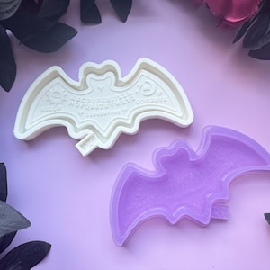 May include: Two silicone molds in the shape of a bat. The top mold is white and features a raised design of a Ouija board. The bottom mold is purple and has a smooth surface.