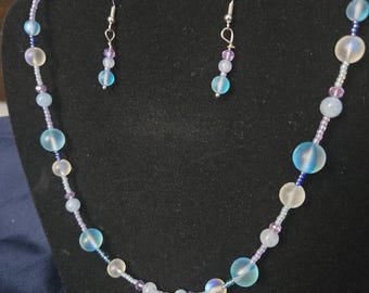 May include: A necklace and earring set featuring iridescent blue and white beads, interspersed with smaller purple beads. The necklace has a silver starfish charm. The earrings have a similar design. The jewelry is displayed on a black mannequin.
