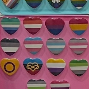 Puede incluir: Una colección de pines en forma de corazón con varios diseños de banderas del orgullo LGBTQ+. Los pines presentan una gama de colores y patrones, incluyendo rayas de arcoíris, degradados y formas geométricas. Los pines están dispuestos sobre una superficie rosa.