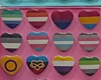 Peut inclure: Une collection d'épingles en forme de cœur avec divers motifs de drapeaux de la fierté LGBTQ+. Les épingles présentent une gamme de couleurs et de motifs, notamment des rayures arc-en-ciel, des dégradés et des formes géométriques. Les épingles sont disposées sur une surface rose.