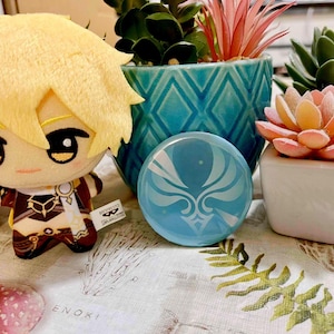Op de afbeelding: Een pluche pop met geel haar en een bruin en gouden outfit zit naast een blauw, cirkelvormig object met een wit ontwerp. Decoratieve potplanten in blauwgroene en witte potten staan ook in de afbeelding.