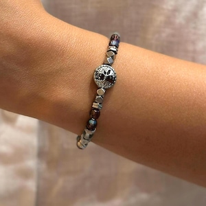 May include: A beaded bracelet featuring a central silver charm with a tree design. The bracelet incorporates a mix of silver, dark red, and iridescent beads. The bracelet is worn on a wrist.