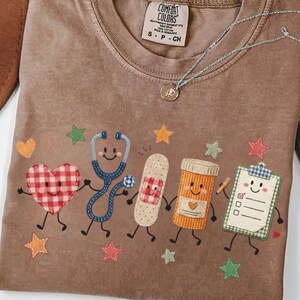 Peut inclure: Un t-shirt marron avec une étiquette Comfort Colors au col, présentant un motif de cinq objets médicaux dessinés : un cœur à carreaux rouges, un stéthoscope bleu, un pansement beige, un flacon de pilules orange et un presse-papiers, avec des étoiles.