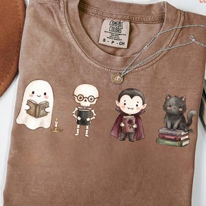 Peut inclure: Un t-shirt marron à manches courtes avec une étiquette Comfort Colors au col. L'illustration représente un fantôme lisant un livre, un squelette à lunettes tenant un livre, un vampire tenant un livre avec une rose et un chat noir sur des livres.