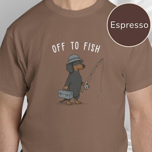 May include: An espresso brown t-shirt featuring a graphic of a black and tan dachshund wearing a grey bucket hat, holding a fishing rod and a grey tackle box, with the white text "OFF TO FISH" printed above the dog illustration.