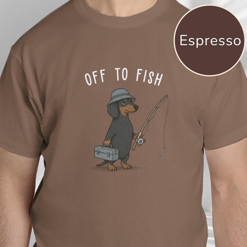 May include: An espresso brown t-shirt featuring a graphic of a black and tan dachshund wearing a grey bucket hat, holding a fishing rod and a grey tackle box, with the white text "OFF TO FISH" printed above the dog illustration.