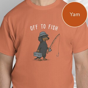 May include: A yam-colored t-shirt featuring a graphic of a black and tan dachshund wearing a grey bucket hat. The dog holds a fishing rod and a grey tackle box. Above the dog, the text reads OFF TO FISH in white capital letters.