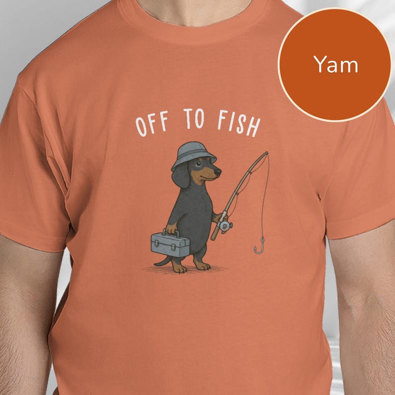 May include: A yam-colored t-shirt featuring a graphic of a black and tan dachshund wearing a grey bucket hat. The dog holds a fishing rod and a grey tackle box. Above the dog, the text reads OFF TO FISH in white capital letters.