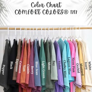 May include: A color chart for Comfort Colors 1717 t-shirts featuring a row of shirts on white hangers. The colors labeled are Pepper, Black, Espresso, Crimson, Yam, Cumin, Berry, Blue Jean, Light Green, Moss, Bay, Violet, Chalky Mint, Lagoon Blue, Neon Pink, Orchid, Mustard, Ivory, and White.