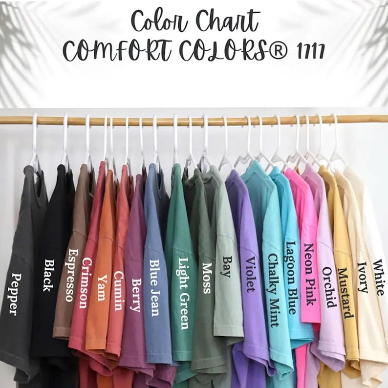 May include: A color chart for Comfort Colors 1717 t-shirts featuring a row of shirts on white hangers. The colors labeled are Pepper, Black, Espresso, Crimson, Yam, Cumin, Berry, Blue Jean, Light Green, Moss, Bay, Violet, Chalky Mint, Lagoon Blue, Neon Pink, Orchid, Mustard, Ivory, and White.