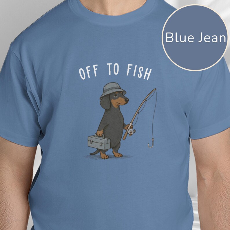 May include: A blue jean colored t-shirt featuring a graphic of a black and tan dachshund wearing a bucket hat, holding a fishing rod and a tackle box. The text "OFF TO FISH" is printed in white above the dog illustration on the front center.