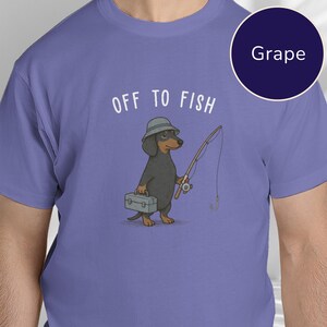 May include: A grape-colored t-shirt featuring a graphic of a black and tan dachshund wearing a grey bucket hat. The dog holds a fishing rod and a grey tackle box. Above the dog, the text reads OFF TO FISH in white, sans-serif capital letters.