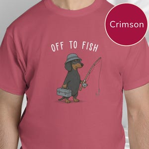 May include: A crimson t-shirt featuring a graphic of a black and tan dachshund wearing a grey bucket hat, holding a fishing rod and a grey tackle box. Above the dog, the text reads OFF TO FISH in white capital letters. The shirt is solid crimson.