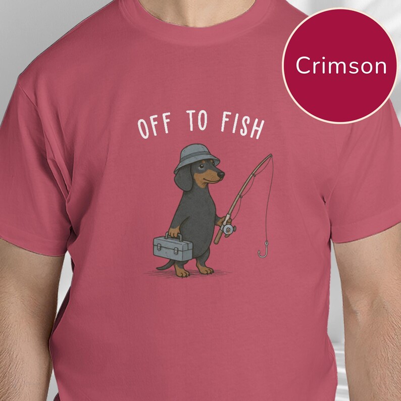 May include: A crimson t-shirt featuring a graphic of a black and tan dachshund wearing a grey bucket hat, holding a fishing rod and a grey tackle box. Above the dog, the text reads OFF TO FISH in white capital letters. The shirt is solid crimson.