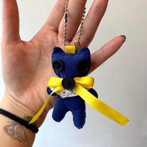 May include: A handmade blue felt cat keychain with black button eyes and a yellow ribbon bow. The cat has a white lace collar and a silver chain for attaching to keys or a bag. The keychain is approximately 3 inches tall.