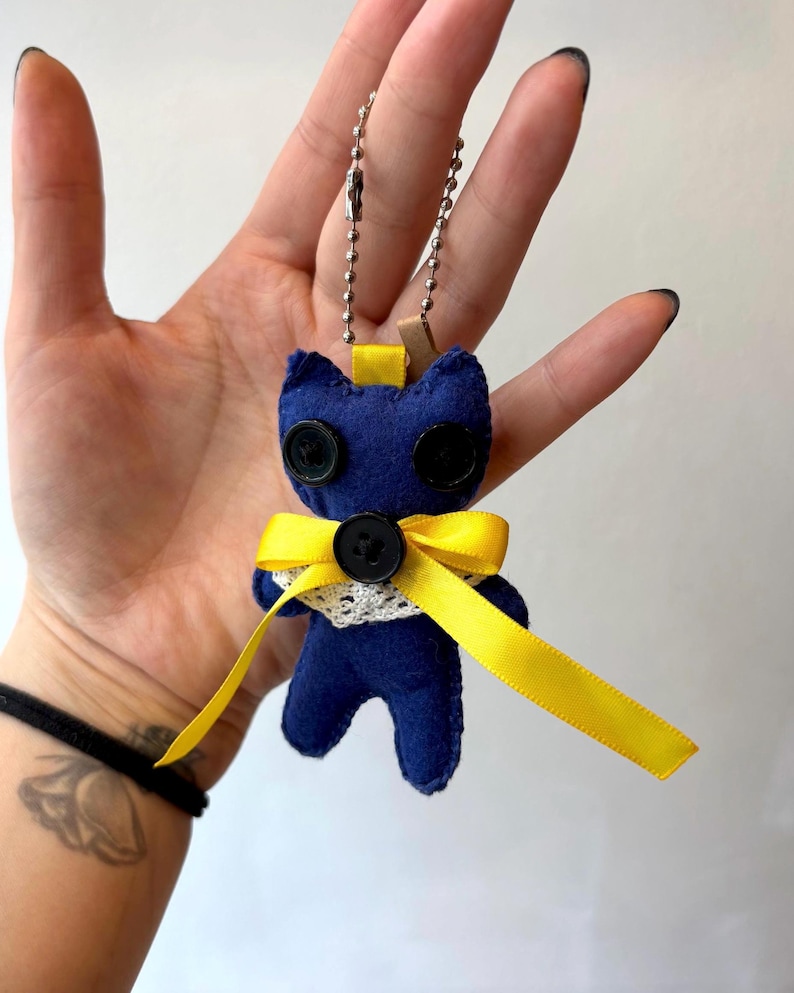 May include: A handmade blue felt cat keychain with black button eyes and a yellow ribbon bow. The cat has a white lace collar and a silver chain for attaching to keys or a bag. The keychain is approximately 3 inches tall.