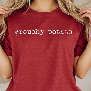 May include: A maroon t-shirt with the words "grouchy potato" printed in white. The shirt is made of a soft material and has a crew neck. The text is centered on the chest.