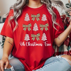 May include: A red t-shirt featuring a festive design with Christmas trees and bows. The shirt has a white text that says "Oh Christmas Tree". The design includes white dotted Christmas trees and green and red bows.