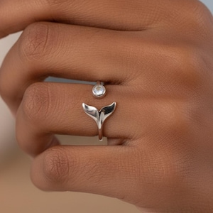 Könnte beinhalten: Ein silberner Ring mit einem Walflossendesign und einem kleinen, klaren Edelstein. Der Schwanz des Rings umschließt den Finger, mit dem Edelstein darüber. Der Ring wird auf einer Hand gezeigt.