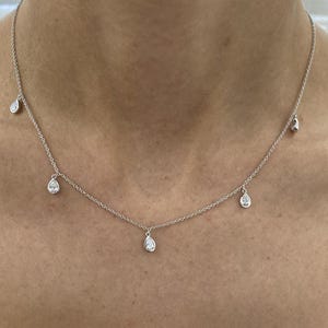 Könnte beinhalten: Eine zarte Silberkette mit mehreren tropfenförmigen Anhängern. Die Anhänger sind gleichmäßig entlang der Kette angeordnet und schaffen ein minimalistisches und elegantes Design. Die Halskette wird vor einem Hautton-Hintergrund gezeigt.