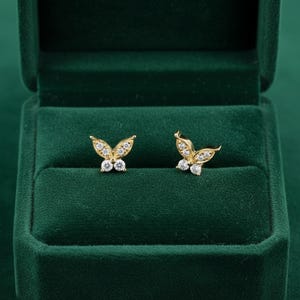 Könnte beinhalten: Ein Paar goldfarbene Schmetterlings-Ohrstecker mit klaren, funkelnden Edelsteinen. Die Ohrringe werden in einer dunkelgrünen Samt-Schmuckschatulle präsentiert. Das Schmetterlingsdesign verleiht dem Schmuck Eleganz.