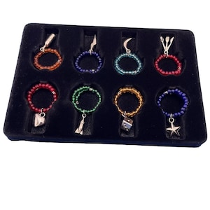 May include: A set of eight beaded wine charms in a black display case. Each charm features a different color of beads and a small silver charm, including a heart, star, and dice. The charms are designed to identify wine glasses.