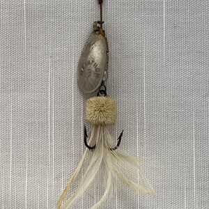 May include: A silver fishing lure with a white feather tail and a white fuzzy body. The lure has a silver blade with the word "Marathon" printed on it.