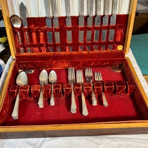 May include: A vintage silverware set in a wooden case with a red velvet interior. The set includes spoons, forks, and knives with decorative handles. The case is open, displaying the arranged utensils.