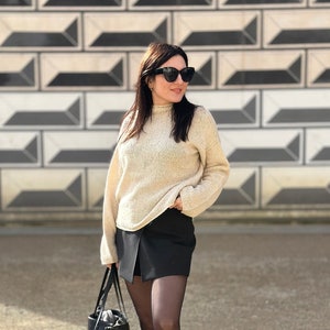 Könnte beinhalten: Eine Frau trägt einen cremefarbenen Pullover, einen schwarzen Rock, schwarze Strumpfhosen und schwarze Combat Boots. Sie trägt eine schwarze Tragetasche und steht vor einer Wand mit einem geometrischen Muster.