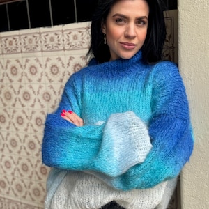 Könnte beinhalten: Ein blau, türkis und weiß farbverlaufender Pullover mit hohem Kragen und langen Ärmeln. Der Pullover hat ein flauschiges, strukturiertes Aussehen. Das Model trägt eine schwarze Jeans.