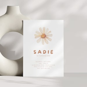 Könnte beinhalten: Eine weiße Einladungskarte mit einem Aquarell-Gänseblümchen-Design und dem Namen "Sadie" in braunen Buchstaben. Der Text "Is turning one" steht unter dem Namen. Zusätzliche Details sind Datum, Uhrzeit und Ort.