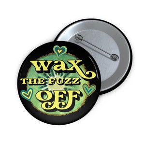 May include: A black button with a green kiwi fruit design and the text "Wax the Fuzz Off" in yellow letters. There are three small hearts in the design.