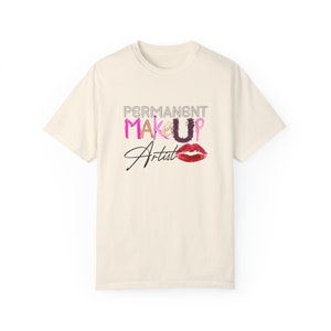 Pode incluir: Uma camiseta branca com o texto "PERMANENT MAKEUP Artist" em letras pretas e rosa. O texto é estilizado com traços de maquiagem coloridos e uma impressão de lábios vermelhos.