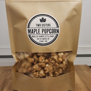 May include: A brown paper bag with a clear window containing maple popcorn. The bag features a circular label with the text "TWO SISTERS MAPLE POPCORN" and "MADE BY HUNGRY LITTLE HANDS IN THE WOODS OF SIDNEY, ME."