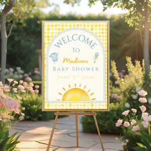 Op de afbeelding: Een babyborrelbord op een houten ezel. Het bord heeft een gele ruitjesrand en bevat de woorden "Welcome to Madisons Baby Shower June 3rd 2026". Het ontwerp bevat een zon, een romper en een rammelaar.