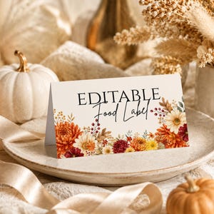 May include: A folded white card with the text EDITABLE Food Label printed in black, featuring a decorative border of autumn-colored flowers including orange, yellow, and deep red chrysanthemums and foliage along the bottom edge of the card.