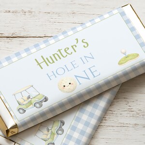 Pode incluir: Embalagem de barra de chocolate com um padrão xadrez vichy azul claro e branco. A embalagem apresenta o texto "Hunter's HOLE IN ONE" em verde e azul, juntamente com ilustrações de uma bola de golfe, um carrinho de golfe e um buraco de golfe.