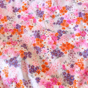 May include: A white fabric featuring a dense, repeating floral pattern. The design consists of small, vibrant flowers in shades of bright pink, deep purple, and vivid orange, interspersed with tiny white blossoms and subtle grey-green foliage.