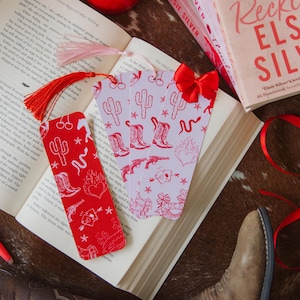May include: Bookmarks with a Western design, showing cacti, cowboy boots, and hearts. A red tassel and bow are attached. A book and a lit candle are also visible. The scene is set on a cowhide-patterned surface.