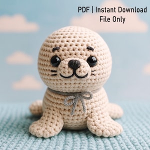 Könnte beinhalten: Ein gehäkeltes Seehund-Plüschtier in Beige mit schwarzen Augen und einer schwarzen Nase. Der Seehund hat eine graue Schleife um den Hals und kleine Flossen. Der Text "PDF | Sofort-Download Nur Datei" befindet sich in der oberen rechten Ecke.