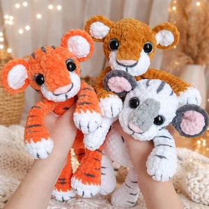 Könnte beinhalten: Drei handgefertigte gehäkelte Tigerbabys in Orange, Braun und Weiß. Der orangefarbene Tiger hat schwarze Streifen, während der weiße Tiger graue Streifen hat. Jedes Baby hat große schwarze Augen, rosa Nasen und weiße Pfoten.