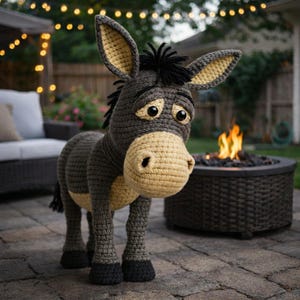 Puede incluir: Una figura de burro de ganchillo hecha a mano en tonos grises, beige y negro. El burro tiene ojos grandes y expresivos y una expresión triste. Las orejas y el hocico son de color beige, y las pezuñas son negras. El burro está sobre un patio de ladrillos.