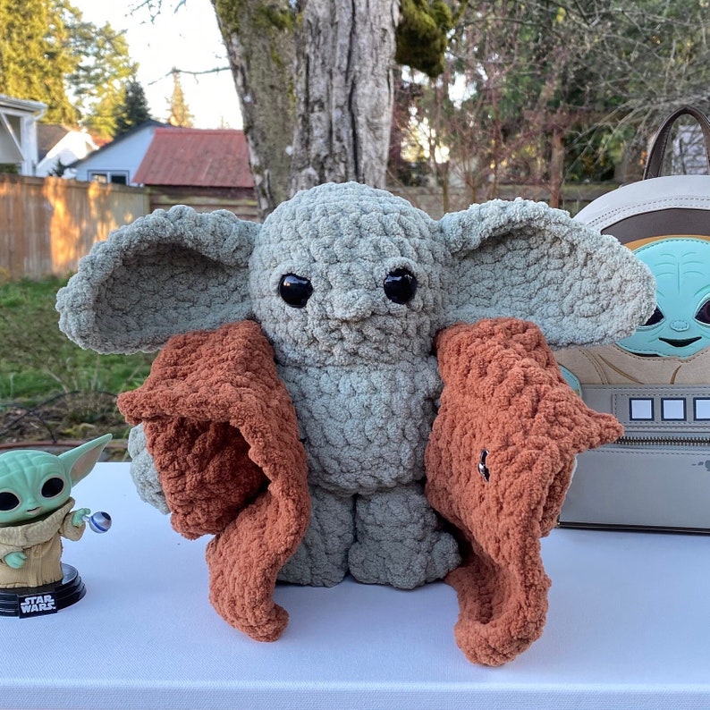 May include: A crocheted plush toy resembling a character from the Star Wars franchise. The toy is green and has large, round eyes. It is wearing a brown crocheted scarf.