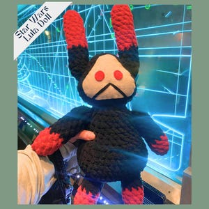 May include: A black and red plush toy shaped like a rabbit with a white face and red eyes. The toy is labeled "Star Wars Lula Doll".