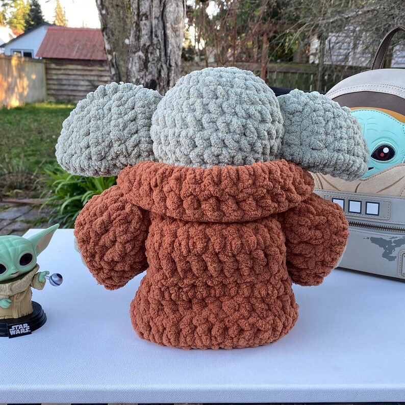 May include: A crocheted plush toy in the shape of a character from the Star Wars franchise. The toy is wearing a brown hooded jacket and has large, floppy ears. The toy is sitting on a white surface.
