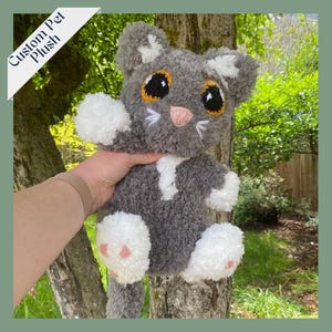 May include: A gray and white custom pet plush toy shaped like a cat. The cat has large, round, brown eyes with yellow pupils, a pink nose, and white paws. The plush toy is being held by a hand in front of a tree.