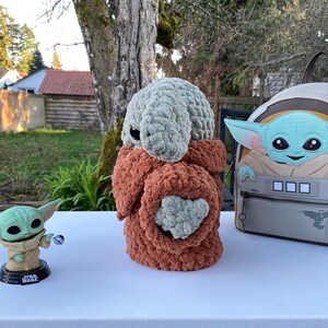 May include: Three crocheted plush toys, two green and one brown, inspired by characters from the Star Wars franchise. The brown plush toy is shaped like a creature with a long snout and is wearing a brown crocheted coat. The green plush toys are shaped like a baby with large ears and are holding a small ball. The toys are sitting on a white table.