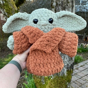May include: A crocheted plush toy of a popular character from the Star Wars franchise. The toy is green and has large ears and big eyes. It is wearing a brown crocheted robe.